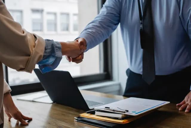 Close-up of two business professionals shaking hands over a wooden office desk with a laptop and contract documents