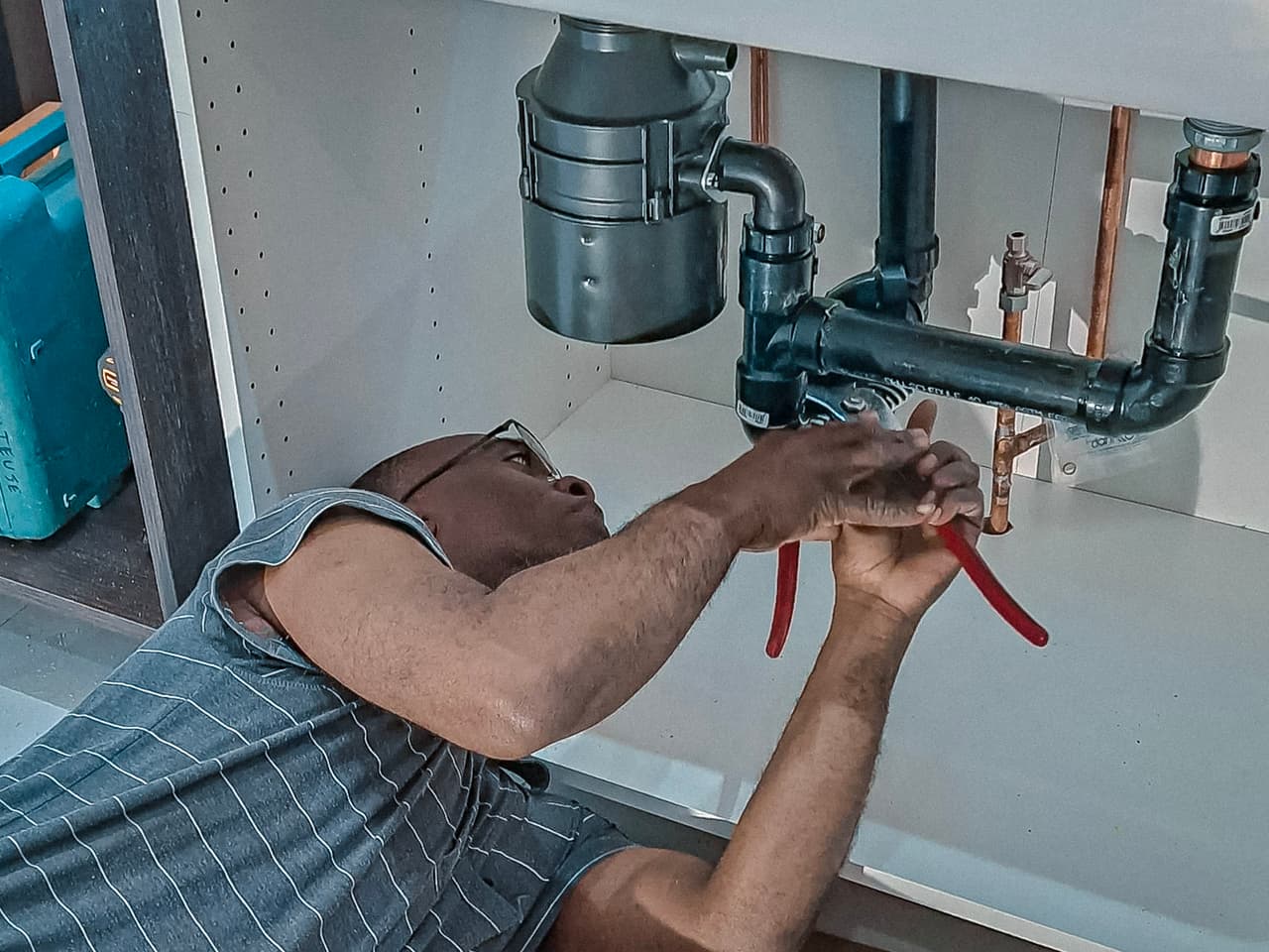 Plumber working under a kitchen sink, repairing pipes with hand tools.
