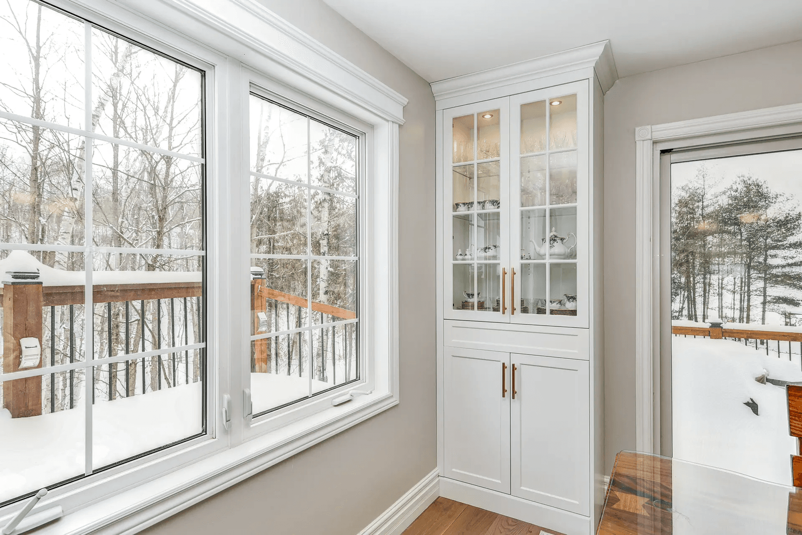 Large white-paned windows offering a panoramic view of a snowy, wooded landscape, allowing an abundance of natural light to flood a bright room, featuring a white display nook and a wooden table nearby.