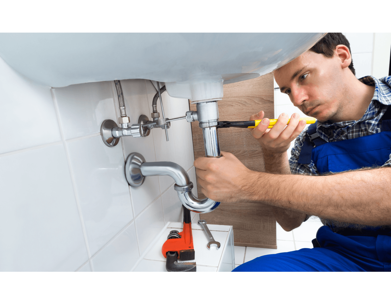 Plumber in blue overalls repairing a sink trap with a wrench in a tiled bathroom.