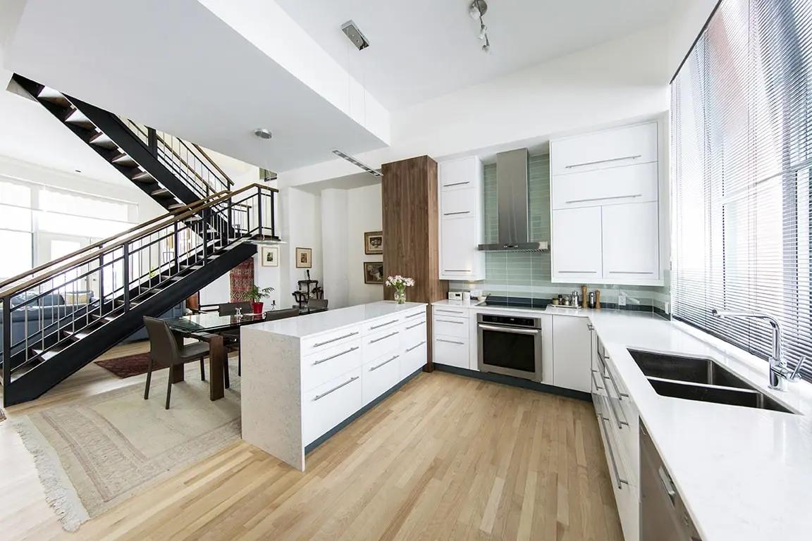 Bright, contemporary kitchen with central island, white quartz worktop and white cabinets. The black metal staircase adds an industrial touch to this space open to the living room. Perfect for lovers of lofts and open spaces.