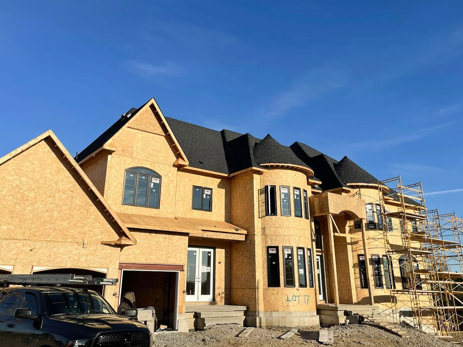 Construction of a large wooden house with black shingle roofing and scaffolding under a blue sky.