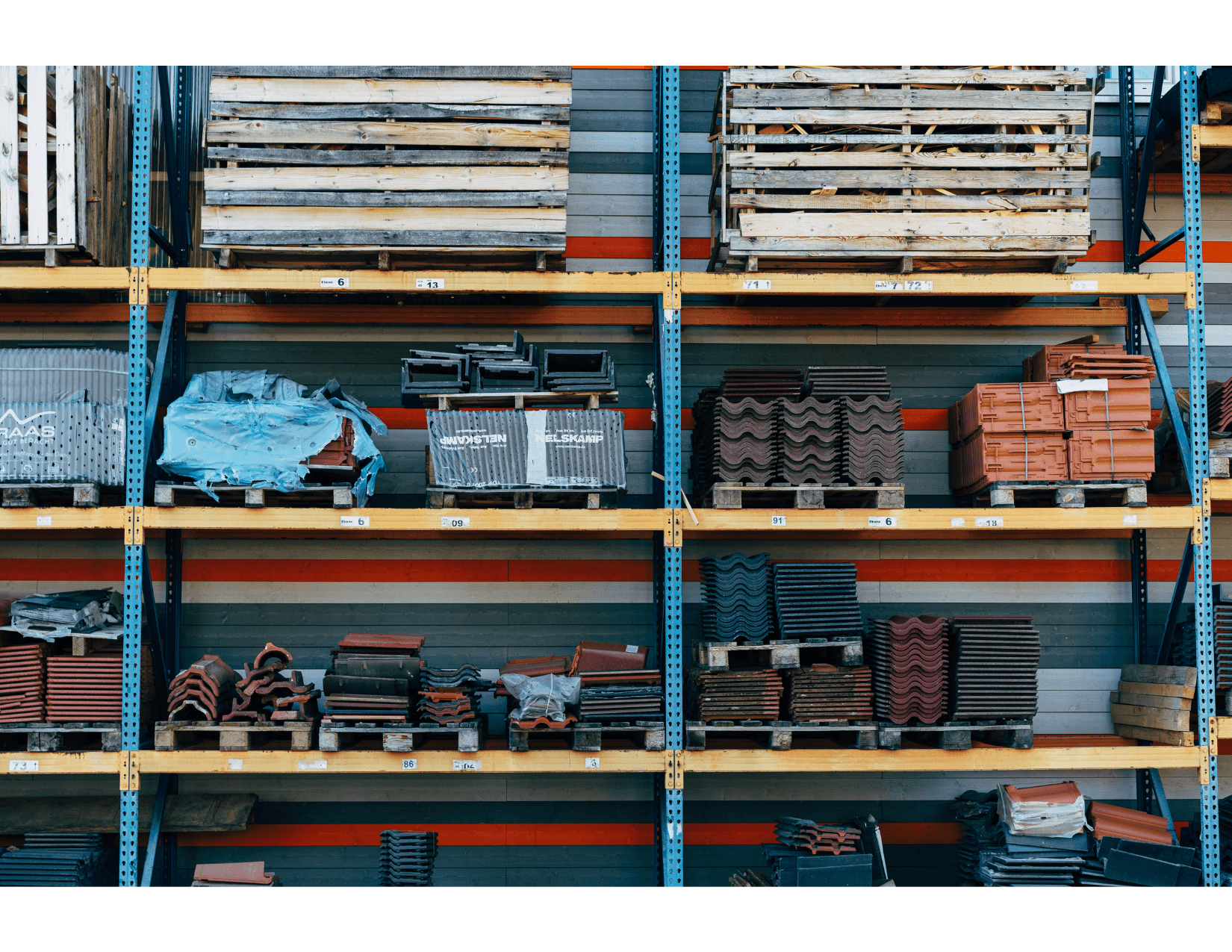 Industrial shelves in a construction materials warehouse, filled with terracotta and metal roof tiles, wooden pallets, and various equipment, organized by type and color.