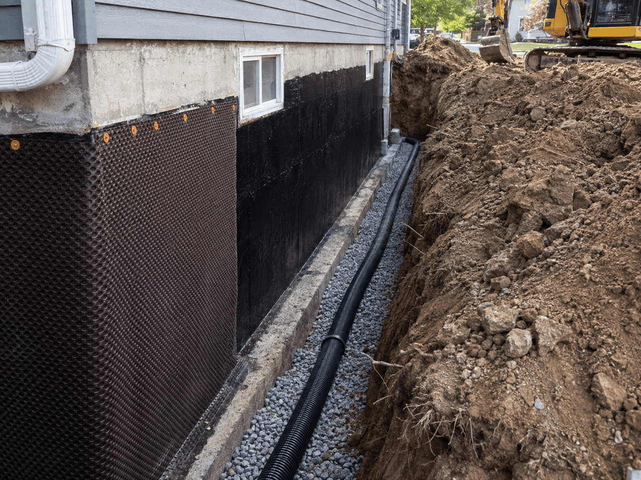 Exterior French drain installation along a home foundation with drainage membrane, perforated pipe, gravel and excavation