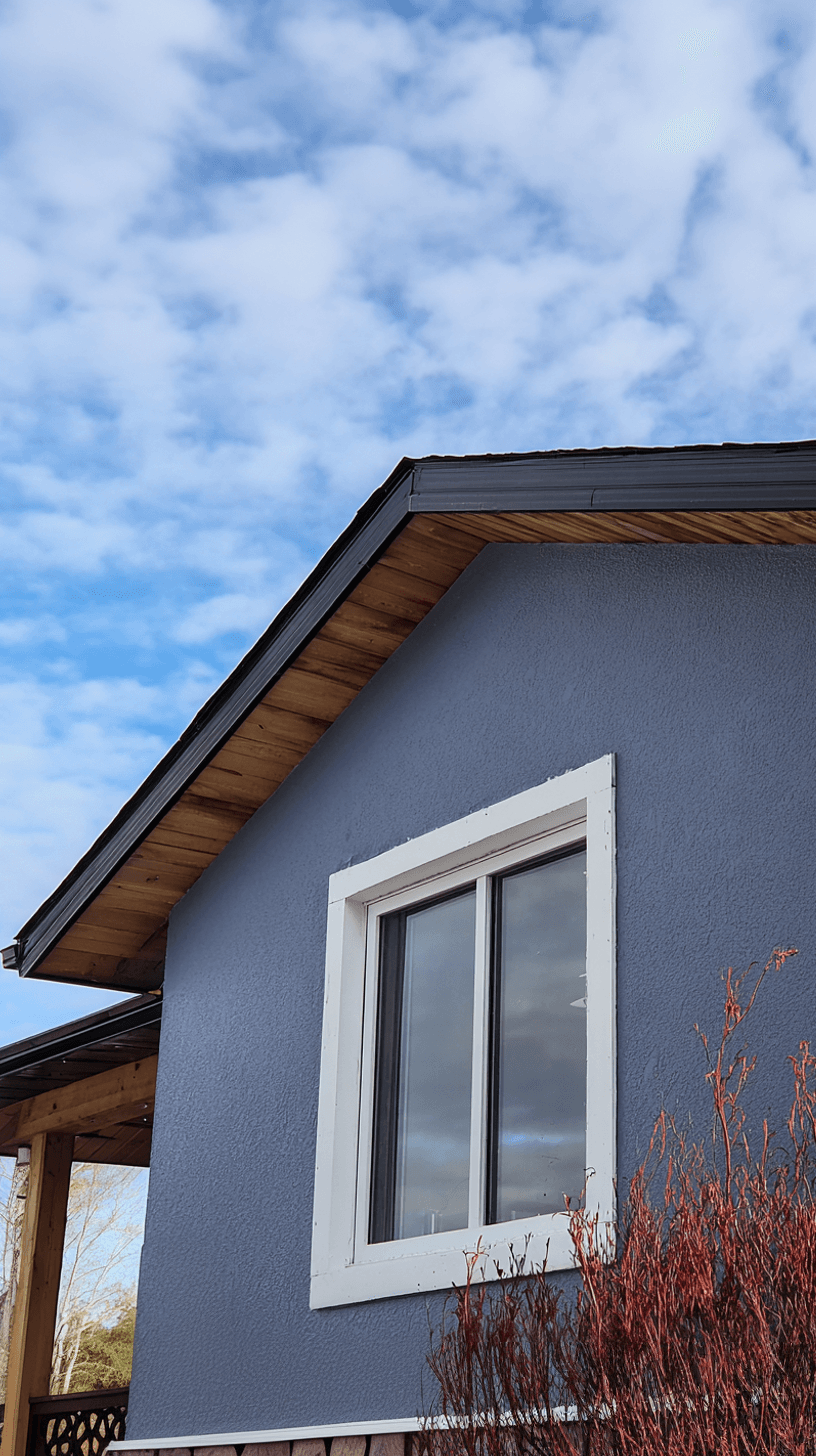 Blue exterior house facade with white window, wood soffits and black roofing under cloudy sky