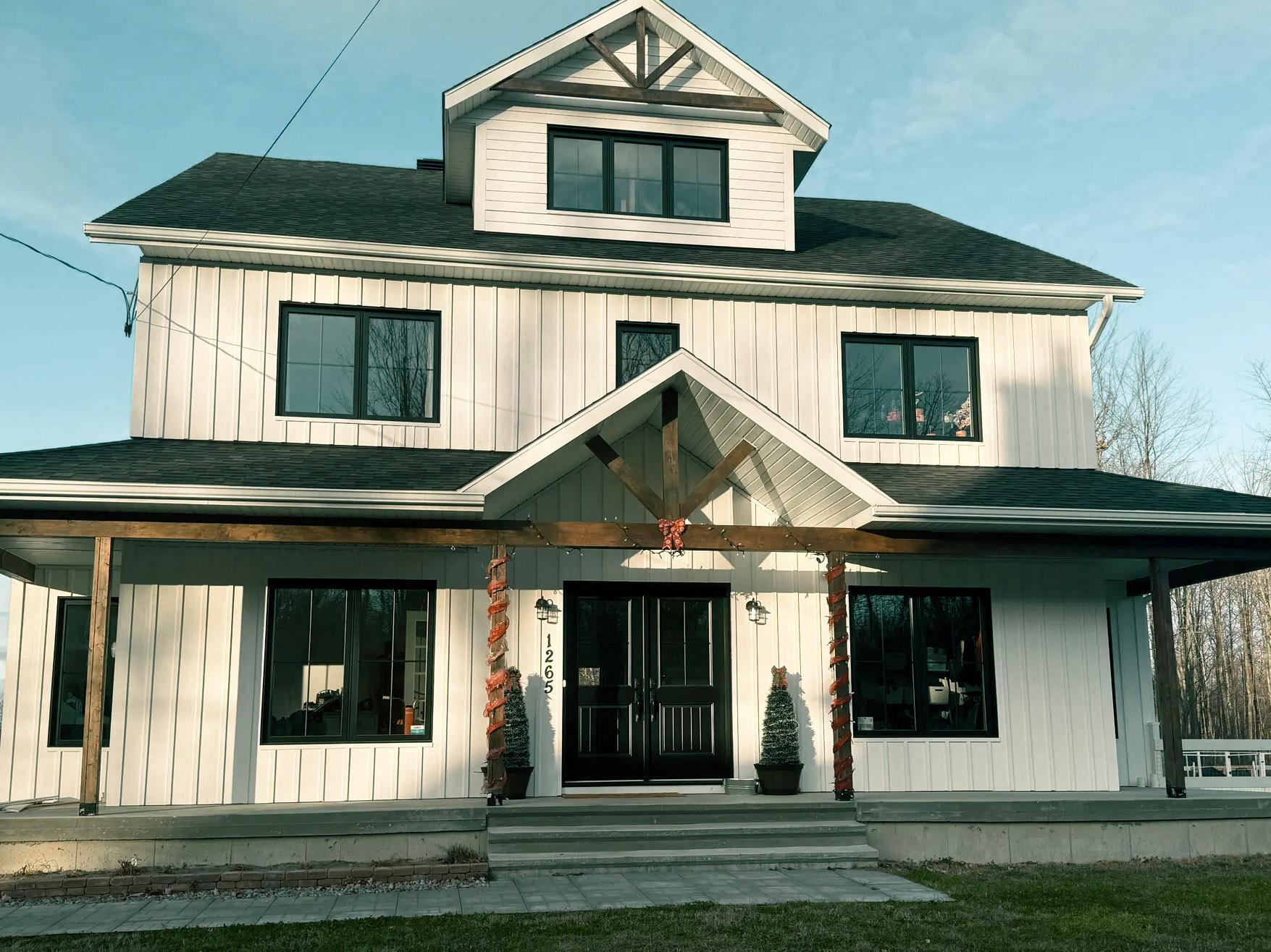 The facade of a modern two-story house with white exterior siding and wooden accents, featuring a welcoming porch adorned with orange garlands for an autumnal ambiance, black windows, and a large double-door entrance.