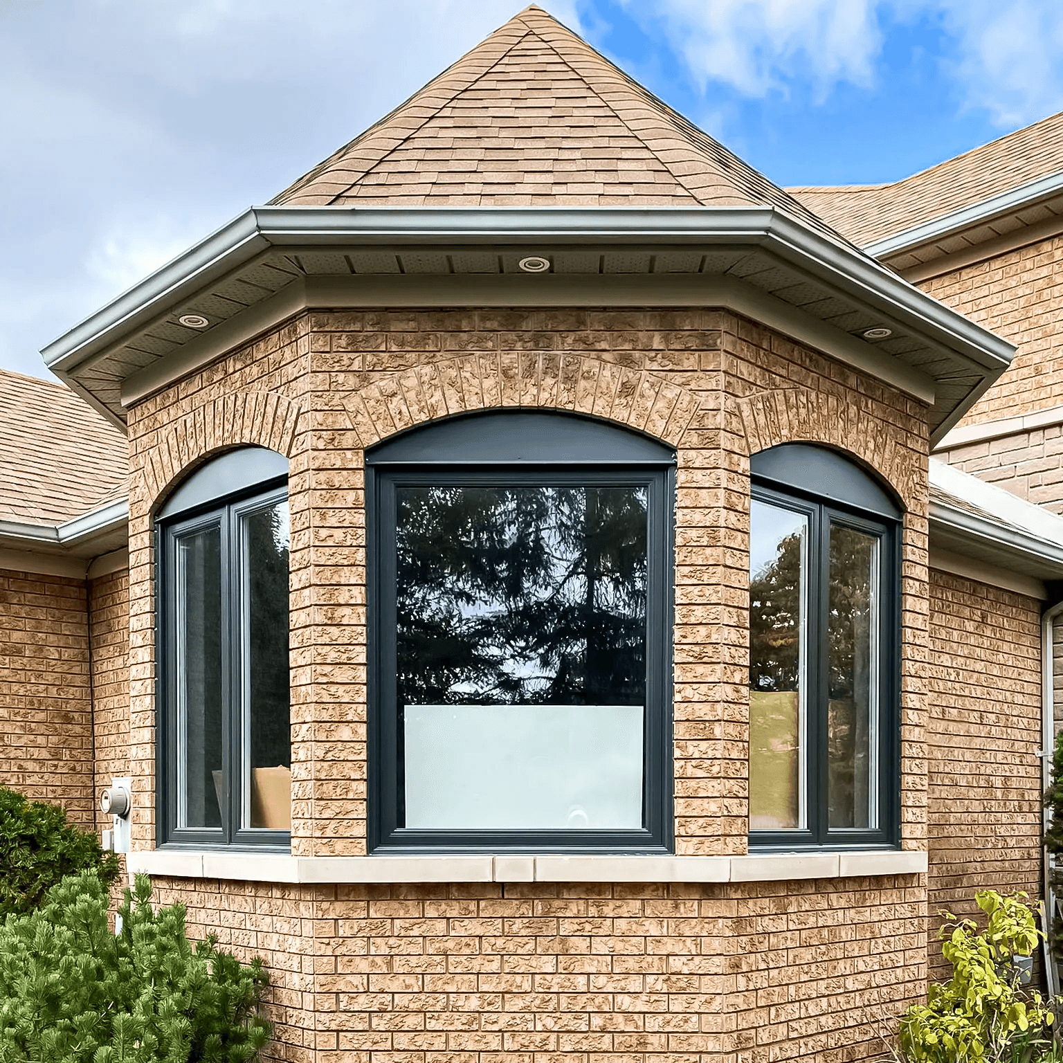 A modern bay window with black frames contrasting against a beige brick facade, highlighting large arched windows and a sloped roof, surrounded by greenery and a partially clear sky.