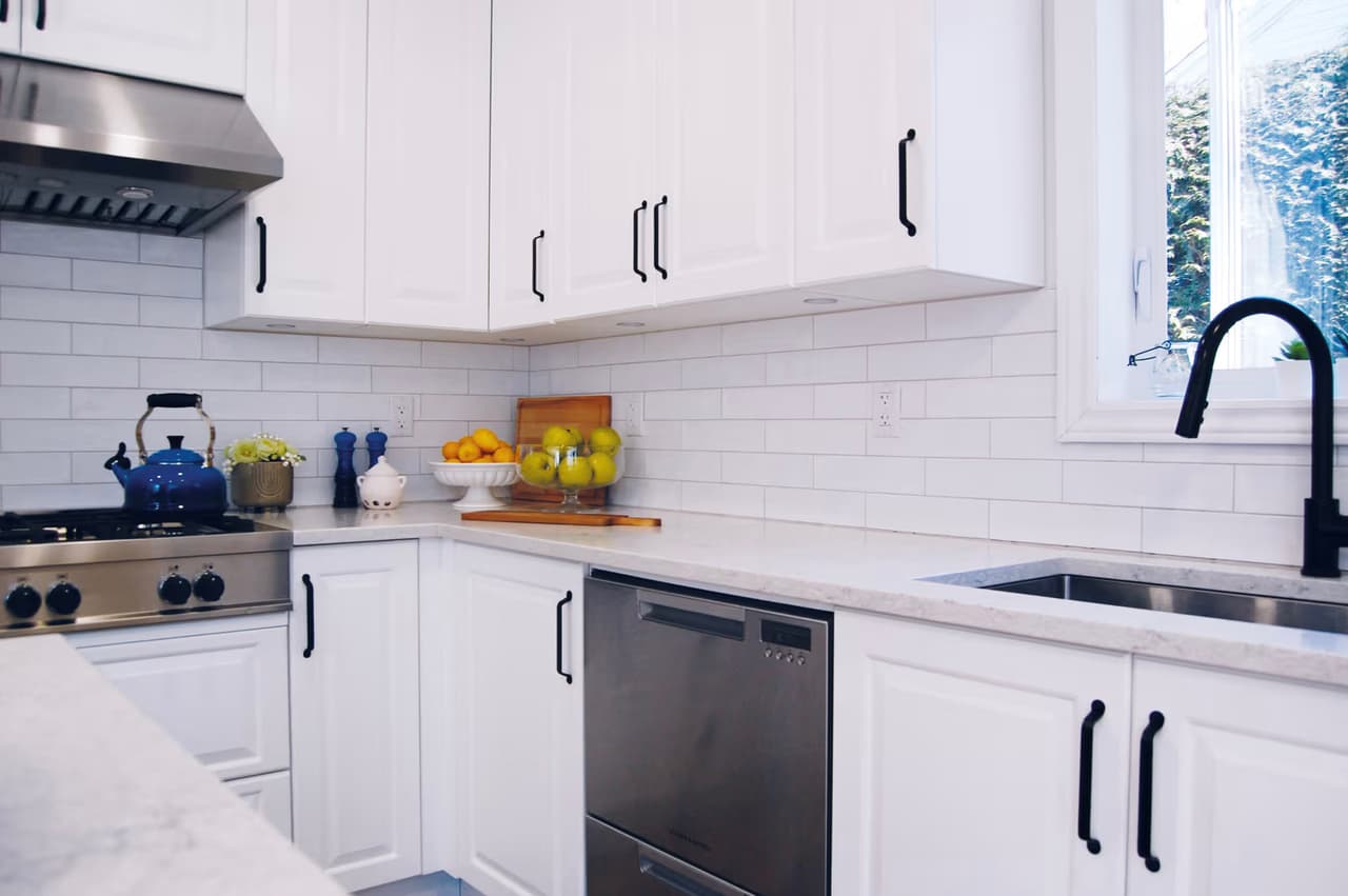 Modern white kitchen with shaker cabinets, black handles, light countertop, subway tile backsplash, stainless steel appliances, and a black faucet beneath the window.