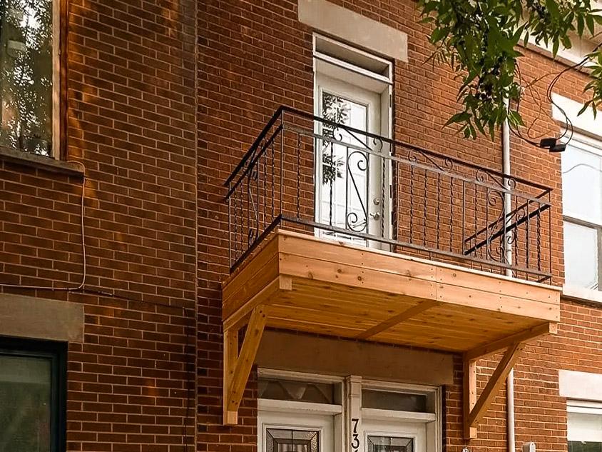 A small wooden balcony with a black wrought iron railing adorned with decorative patterns, located on the upper floor of a red brick house with a white door providing access to the outside, surrounded by an urban setting with trees in the background.