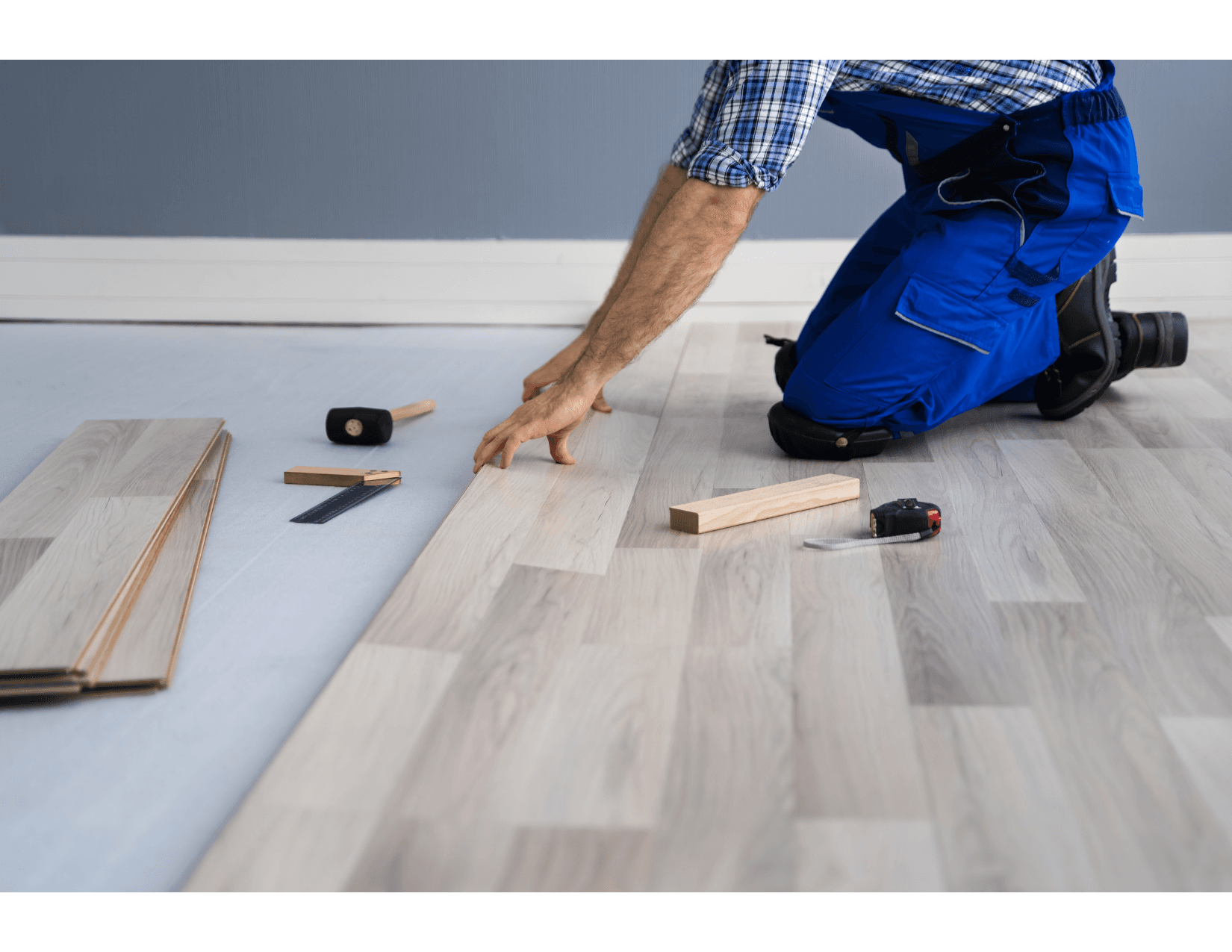 A worker installing new light laminate flooring in a room, kneeling on the ground, surrounded by tools such as a rubber mallet, a ruler, and a measuring tape.