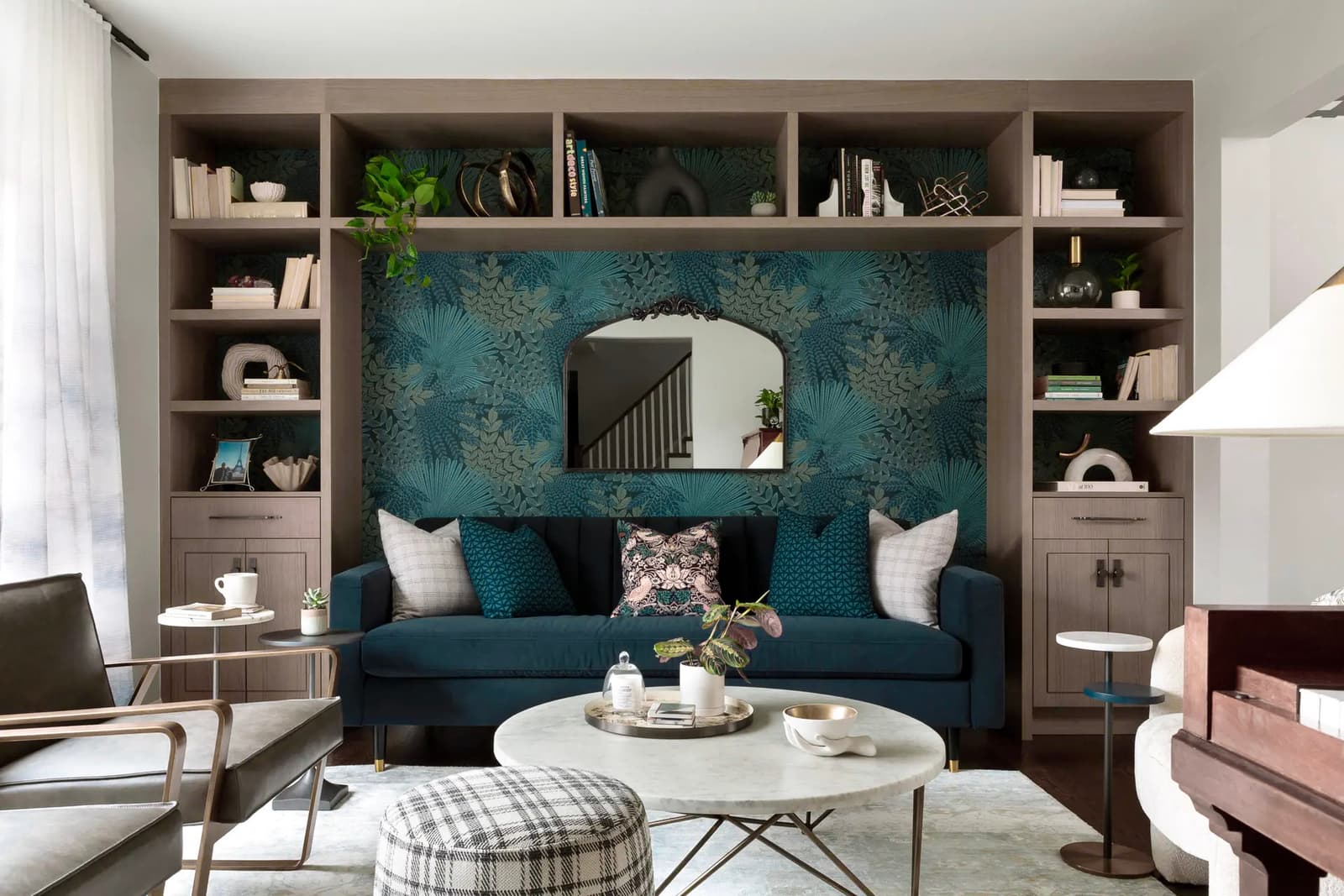 Modern living room with a blue velvet sofa, floral wallpaper, designer mirror, built-in bookshelf, and marble coffee table in a bright and elegant space.