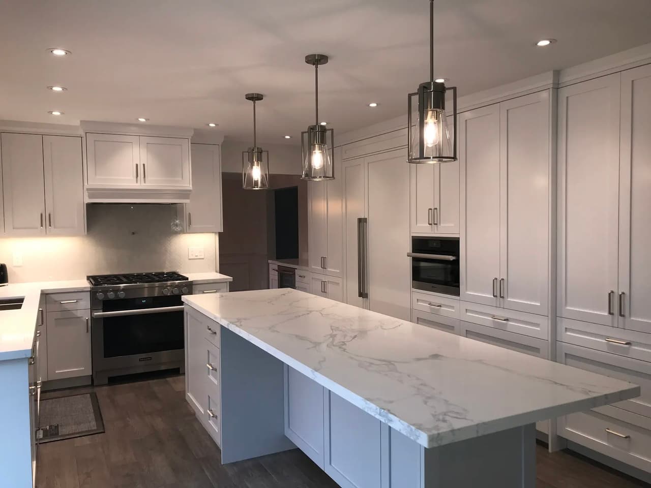 Modern white kitchen with a large central island, marble-look countertop, custom cabinetry, built-in appliances, pendant lights, and recessed lighting.
