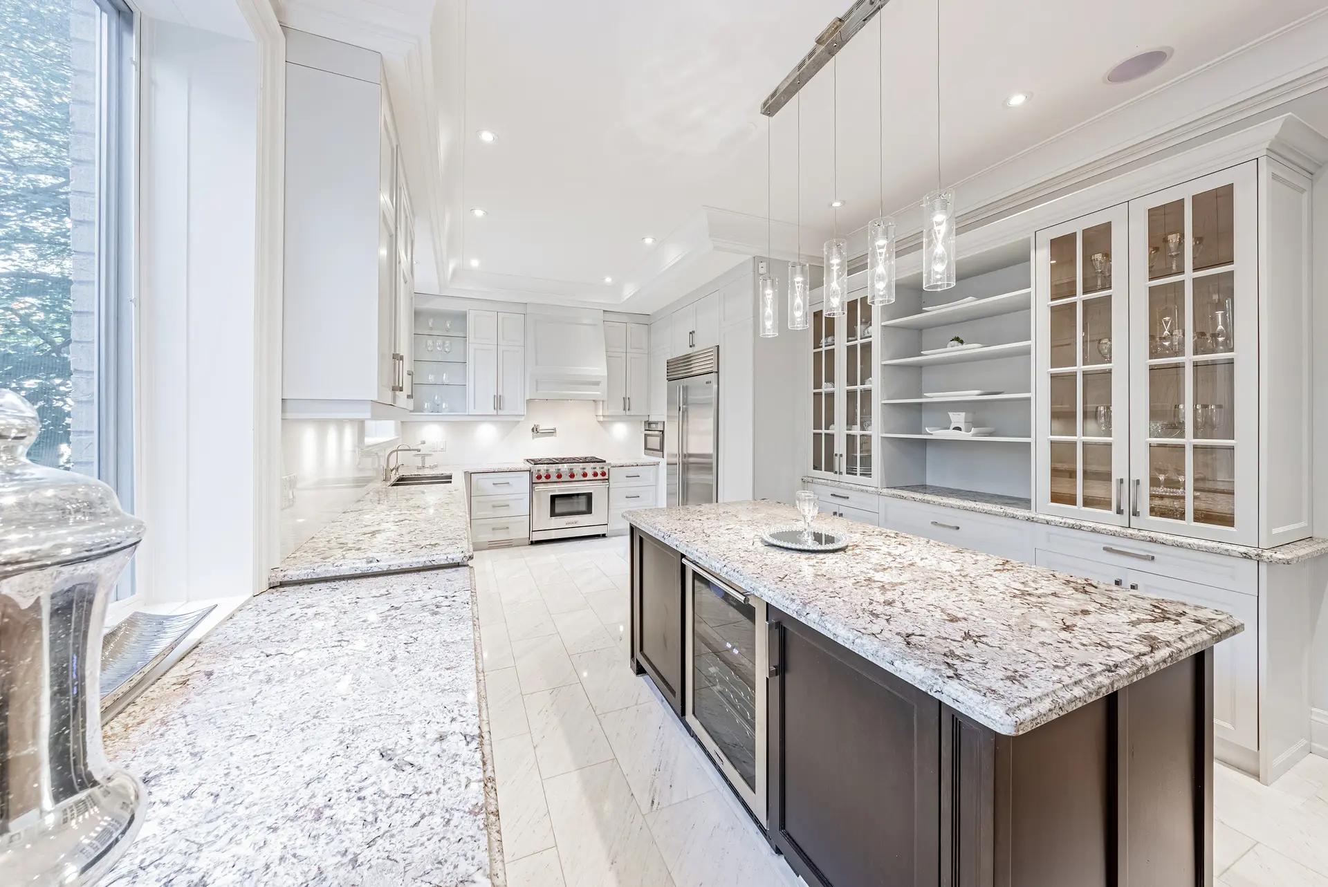 Transitional-looking Montréal kitchen, featuring a spacious central island paired with a granite countertop with veining. The white cabinetry and brushed nickel handles add a touch of elegance to the space. Neutral ceramic flooring creates a harmonious backdrop.