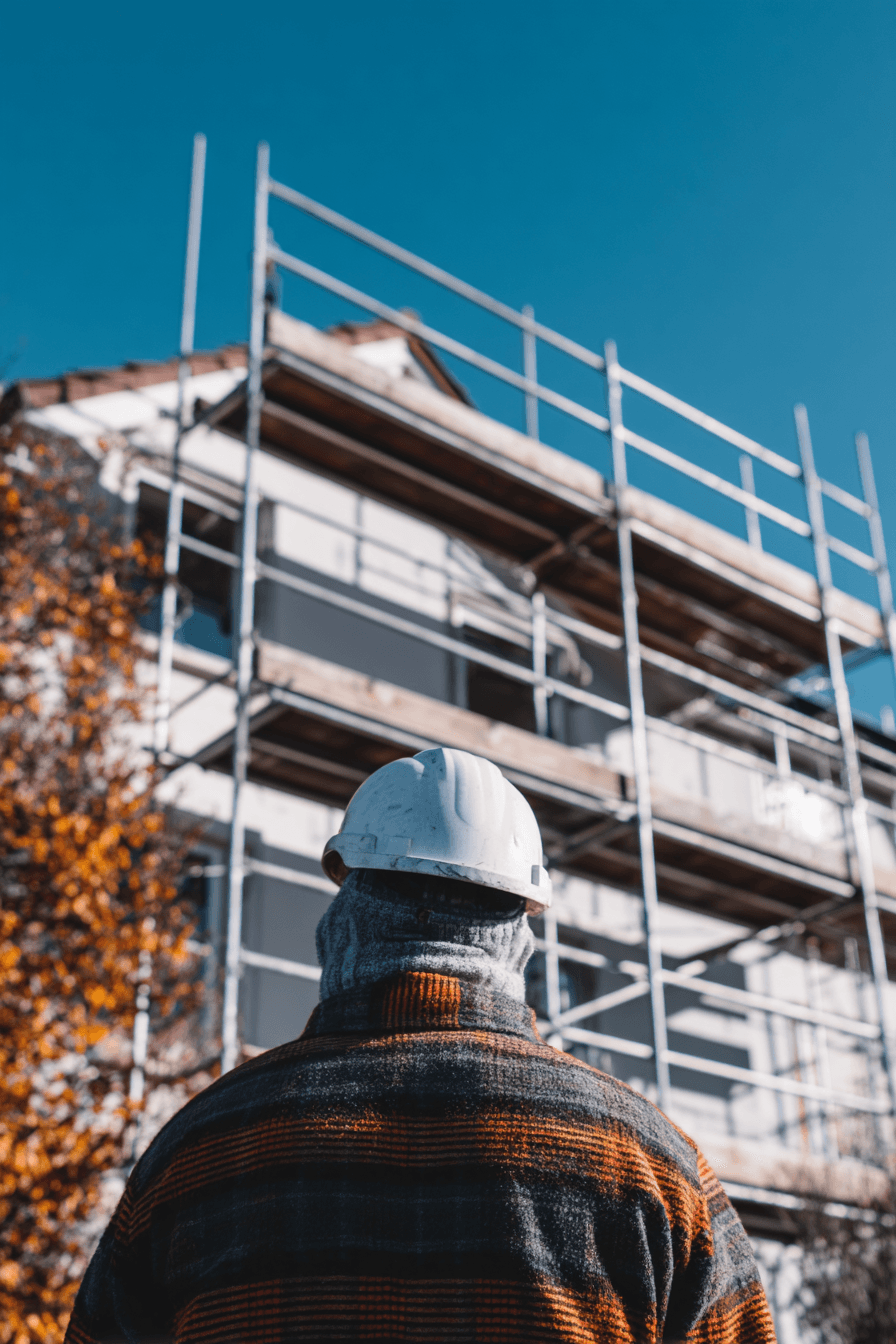 Construction worker wearing a white hard hat looking at a house under exterior renovation with scaffolding under a blue sky