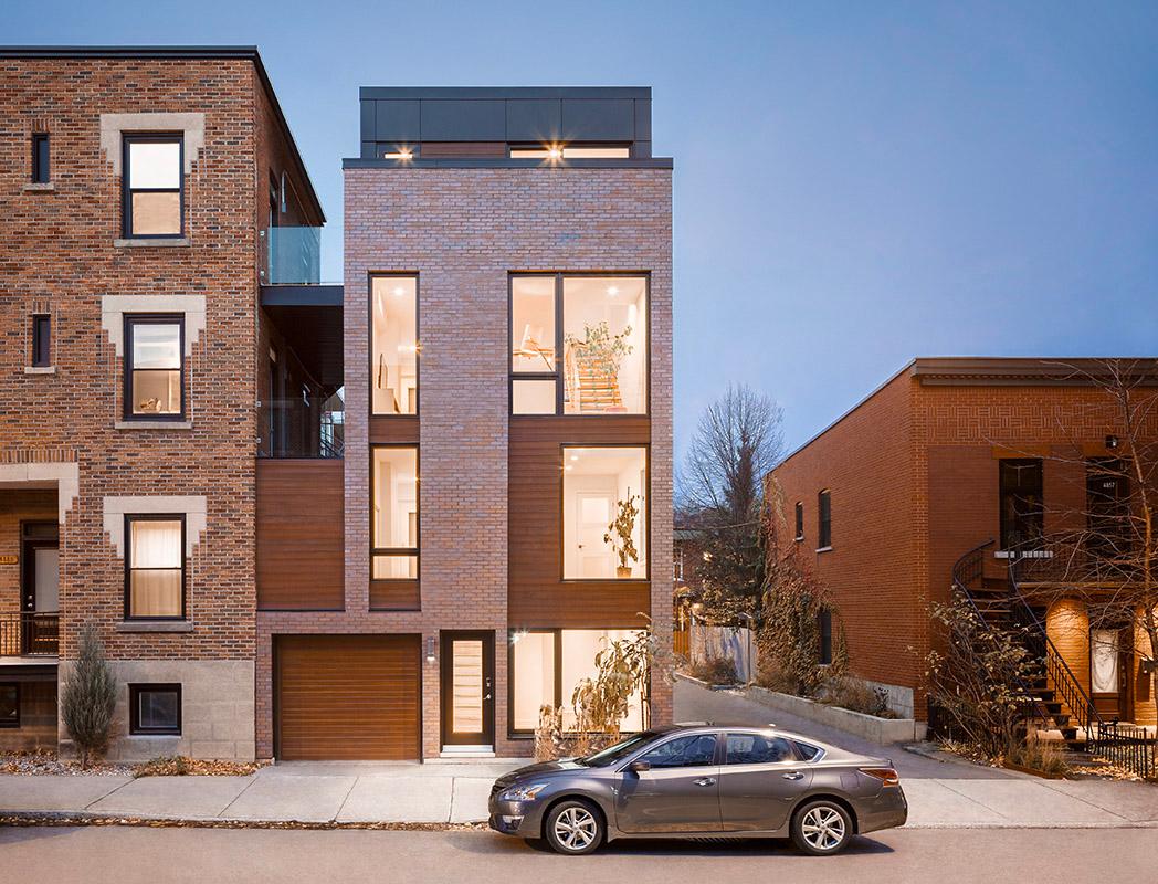 A contemporary house located in Montreal, built with red bricks and wooden panels, featuring large vertical windows that illuminate the interior, a garage on the ground floor, and a sleek design, surrounded by similar urban buildings in a residential neighborhood.