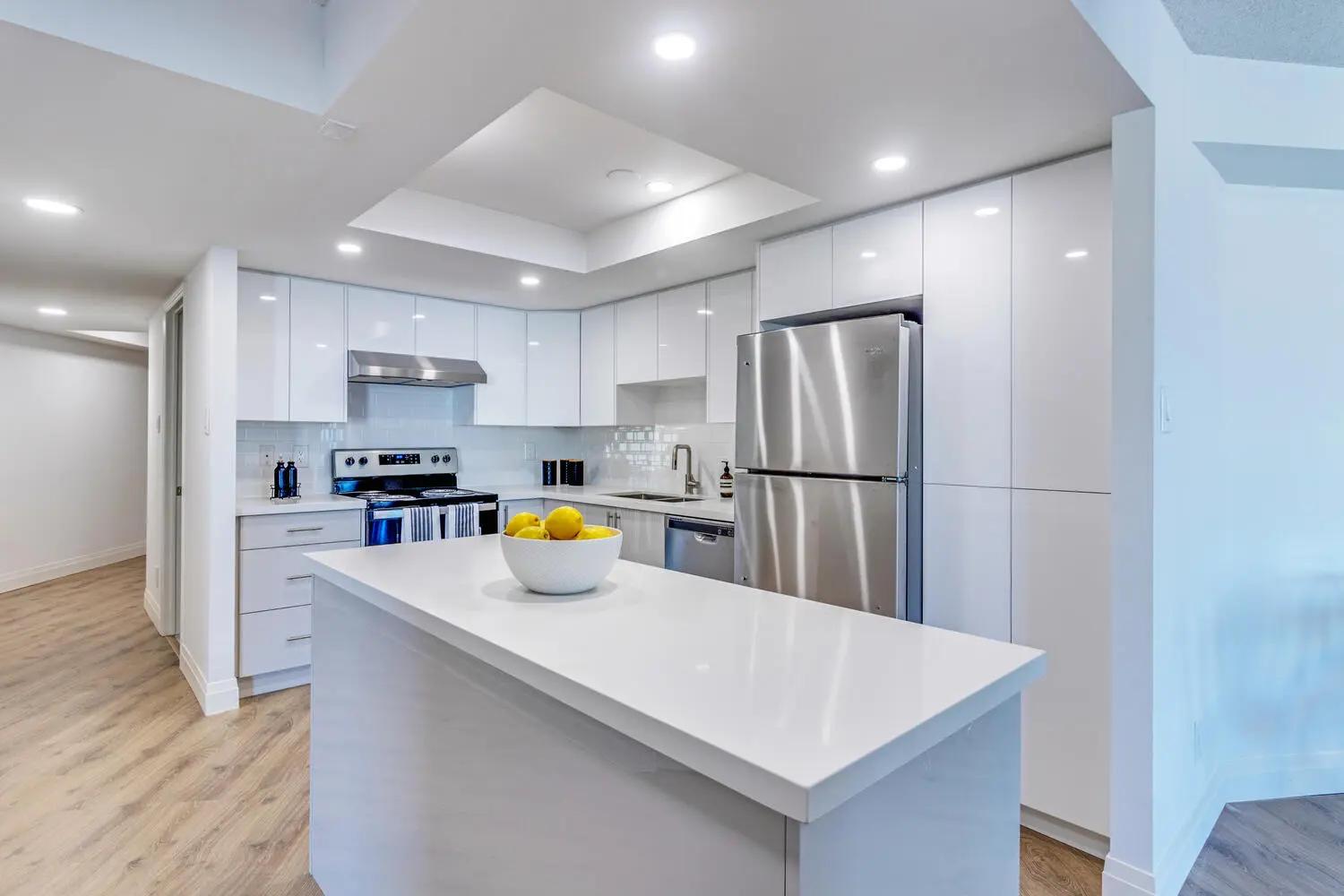 Contemporary-looking Montréal kitchen, featuring a white central island with a quartz countertop. The glossy white cabinets and stainless steel appliances add a hint of elegance to the space, while&nbsp;the light wood flooring and recessed lights make for a bright and inviting setting.