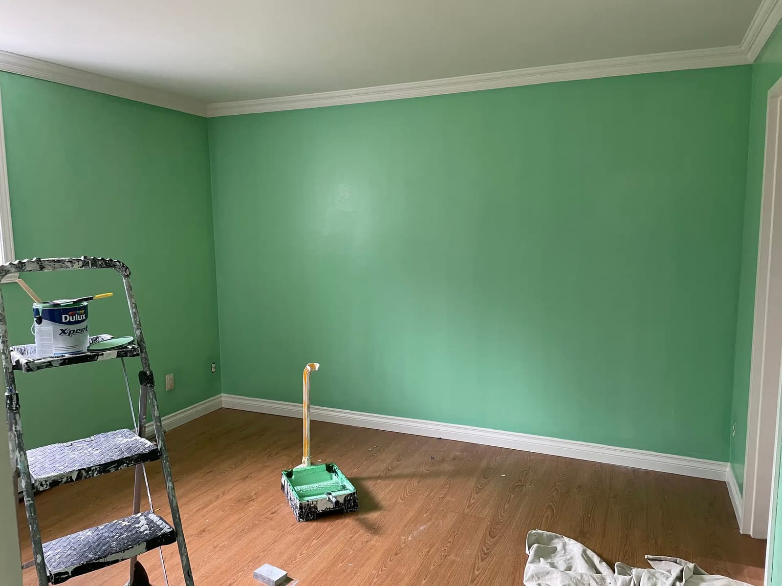 Room under renovation with freshly painted green walls, white moldings, wooden floor, and painting equipment on the ground.