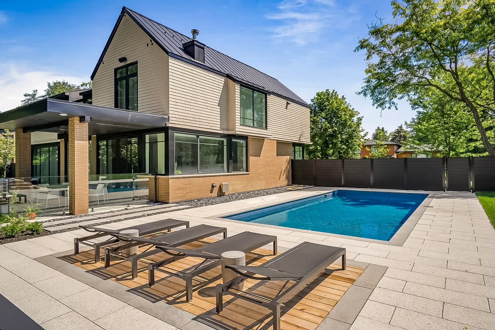 Contemporary house with an inground pool, modern patio, and lounge chairs, surrounded by greenery.