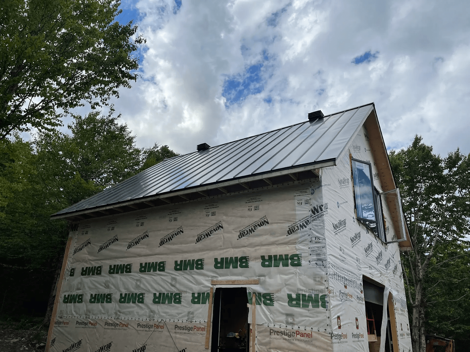 Residential building under construction, with grey metal roof installed. The walls are still covered with protective materials and temporary insulation bearing the 'BMH' and 'PrestigePanel' logos. Set in a natural setting surrounded by trees, the structure is designed with a gabled roof and an upstairs window. The cloudy sky adds an open-air worksite atmosphere.