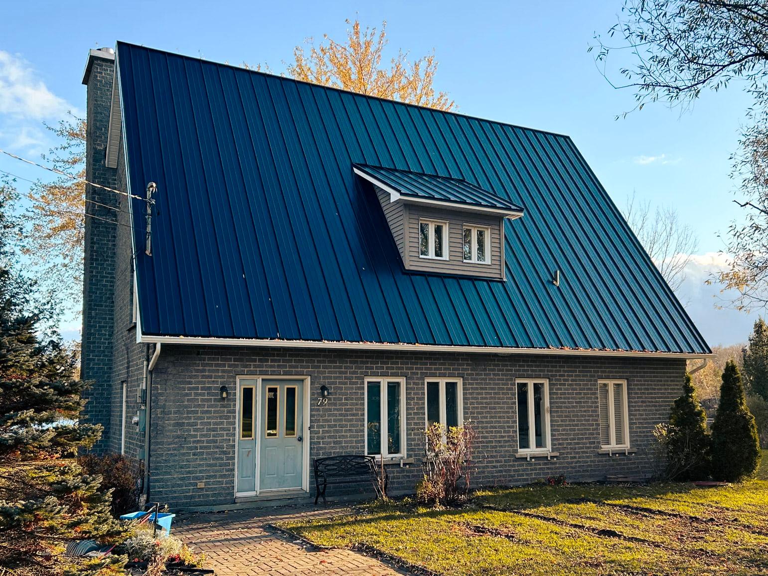 A grey stone house with a steeply pitched dark blue steel roof, featuring a central dormer and large white windows, surrounded by an autumnal landscape with trees and a well-maintained lawn.