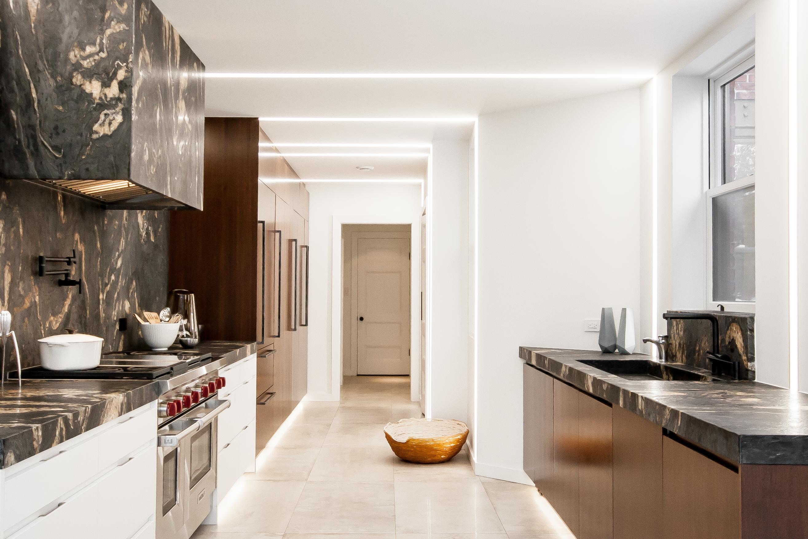 The image showcases a modern and sophisticated kitchen. It features a natural stone backsplash and countertop with dramatic veining, recessed ceiling lighting that enhances the sleek and clean design, and a combination of white and dark wood cabinetry. The layout includes a high-end stainless steel stove and a separate sink area with matching stone surfaces, creating a cohesive and luxurious look. The overall design emphasizes warmth and elegance.