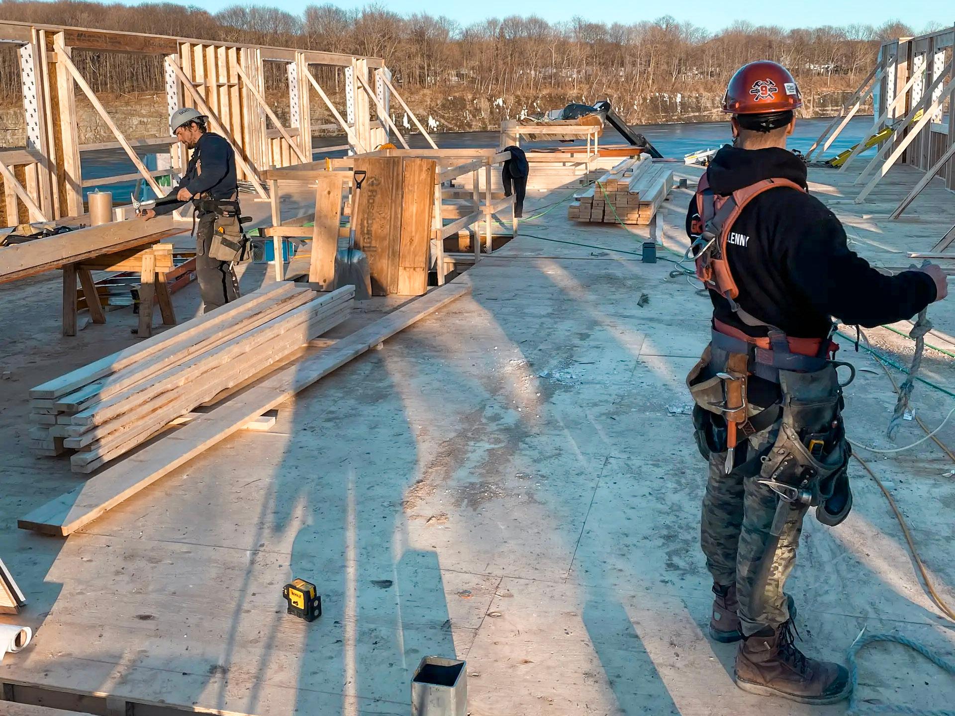 A wooden construction site with several workers wearing safety harnesses and helmets, handling planks and materials under sunny light, in an open environment surrounded by hills.