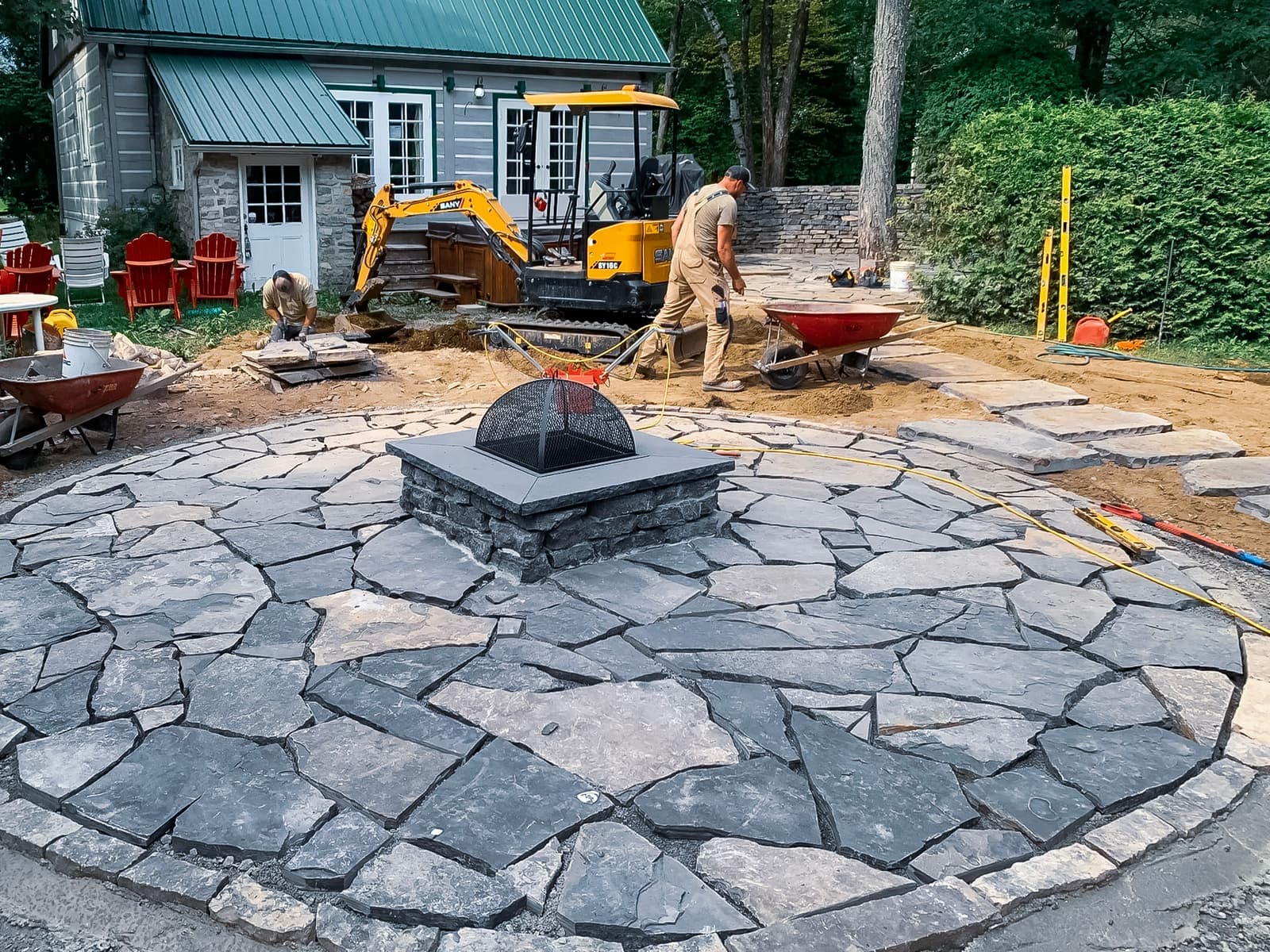 Installation of an outdoor fire pit on a natural stone surface in a landscaping project under construction in front of a wooden and green metal house.