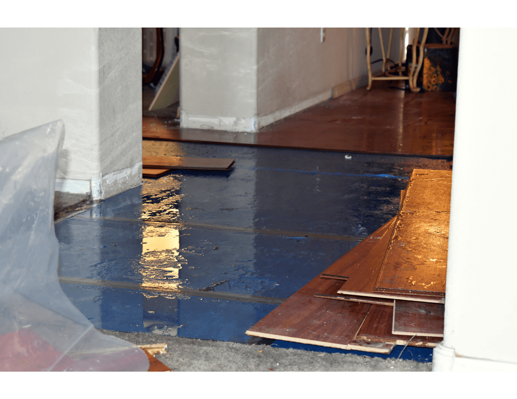 The interior of a house affected by flooding, with stagnant water on the blue floor and stacked wooden planks set aside. The walls and furniture are visible in the background.