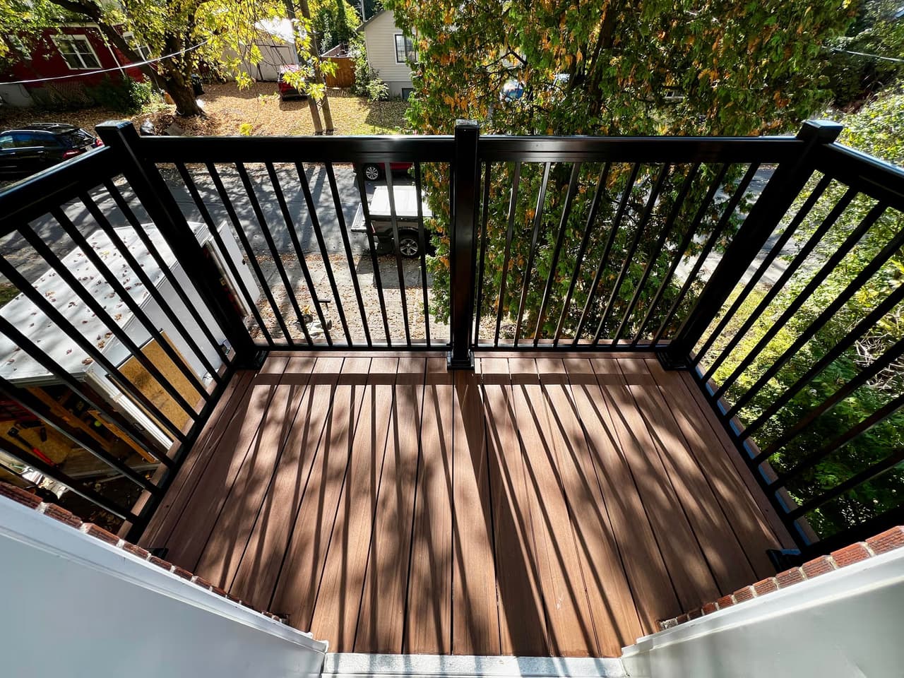 Residential balcony with brown wood flooring, black metal railing and view of trees, street and neighboring houses.