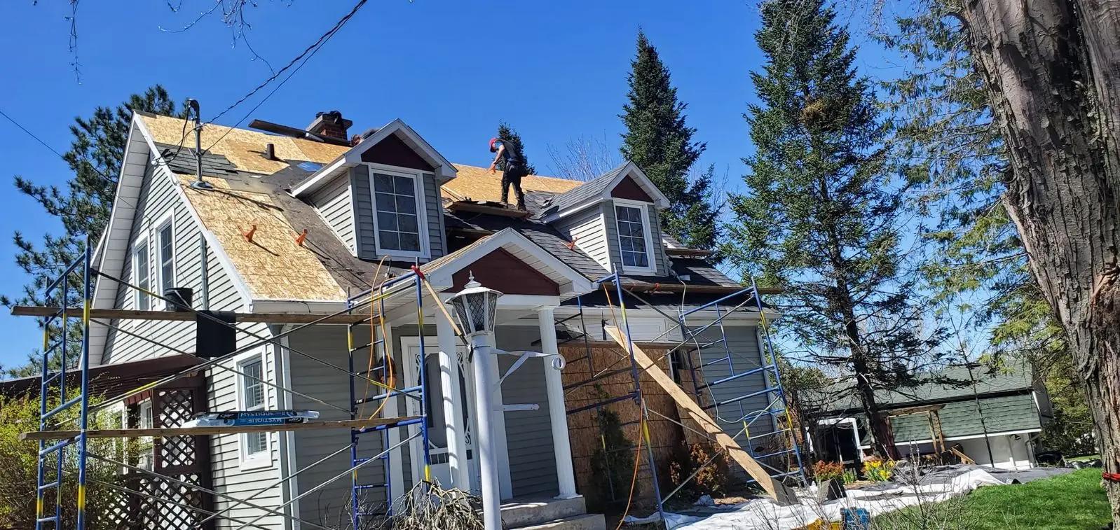 Roof renovation work on two-story house, with workers installing new plywood panels, sourrounded by scaffolding in a wooded residential setting on a sunny day.&nbsp;