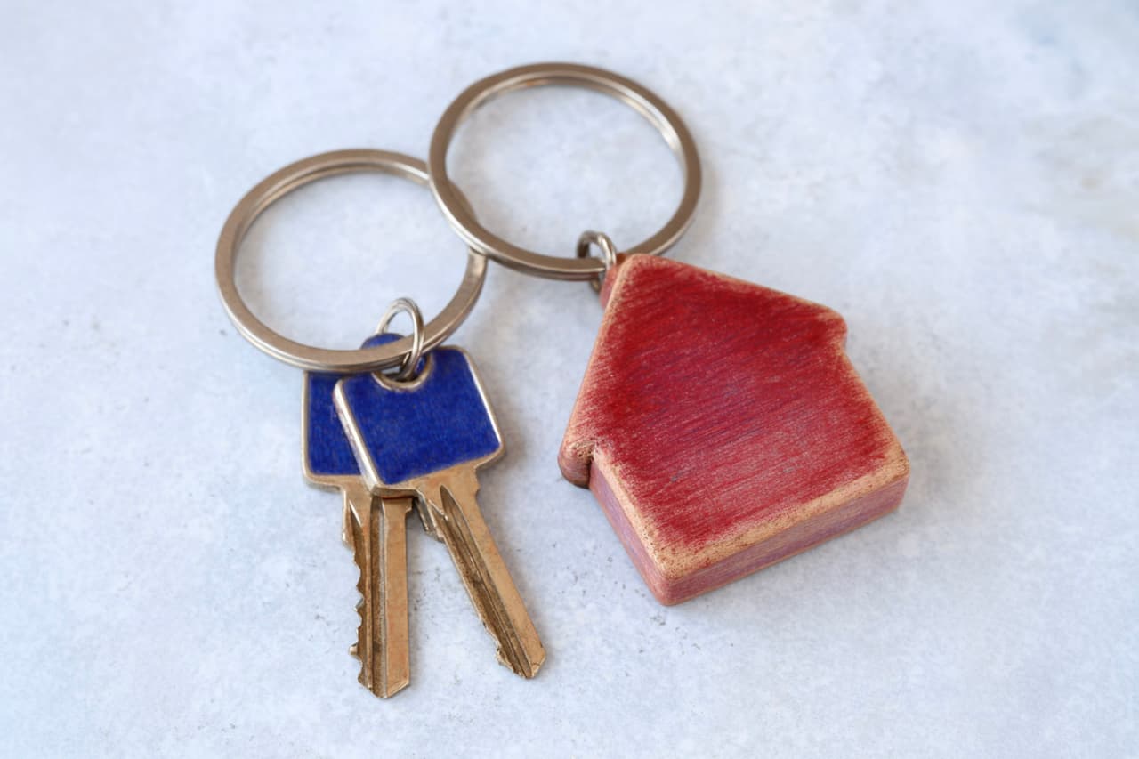 Red house-shaped keychain with metal house keys on a light surface, symbol of property, housing and residential security.