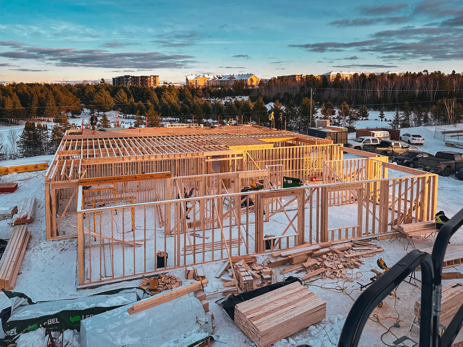 View of a residential construction site with a wooden frame under assembly on a snowy landscape.