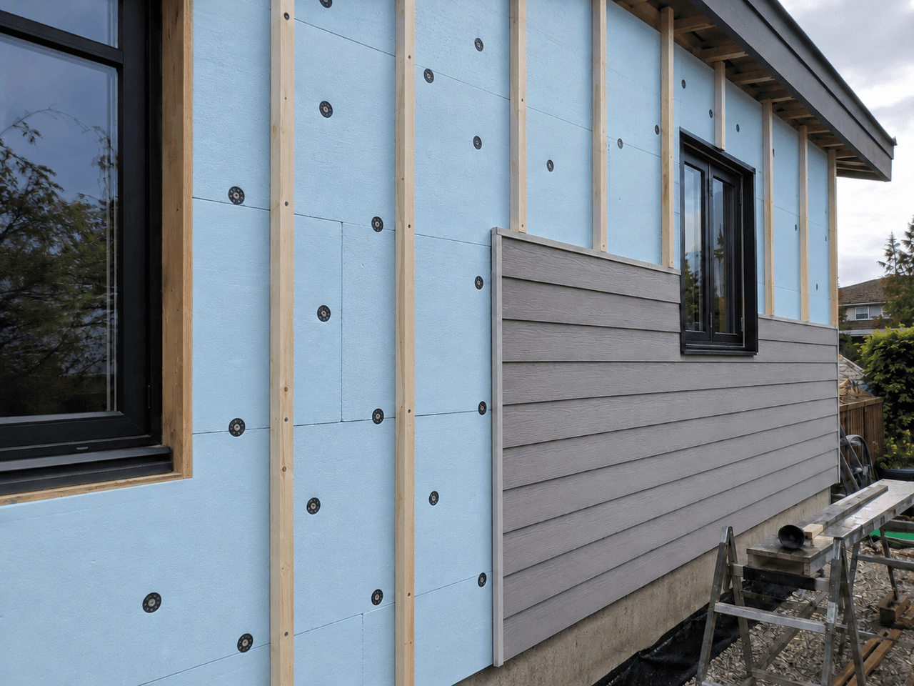 Exterior wall under renovation with blue rigid insulation panels, wood furring strips, black windows and partially installed grey siding