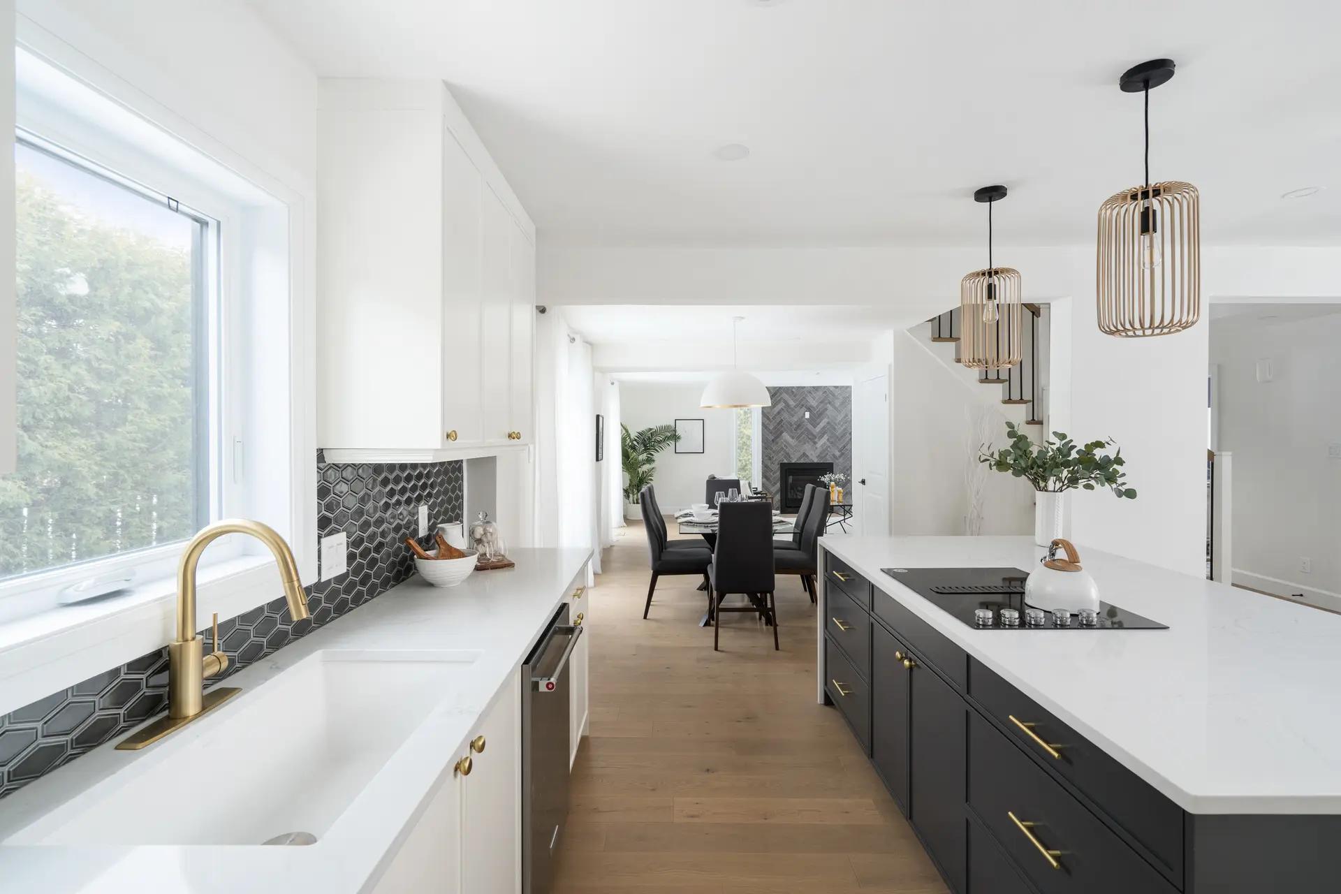 Contemporary-looking Montréal kitchen, featuring a matte black central island with a white quartz countertop. The white shaker cabinets and brass handles add a touch of elegance to the space. The light-hued wooden flooring and suspended light fixtures make for a warm, inviting setting.