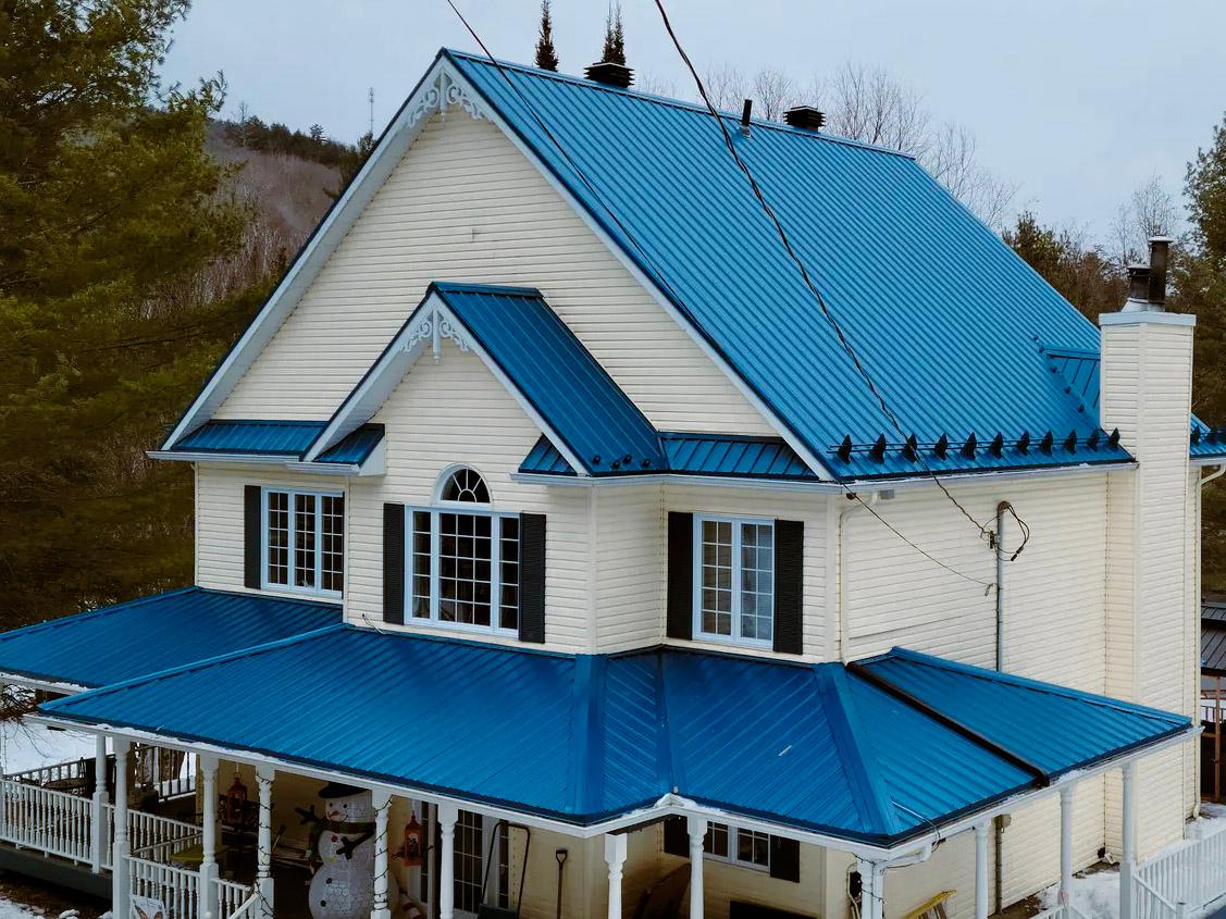 A traditional two-story house with a bright blue steel roof, featuring decorative architectural details, a large central arched window, black shutters, and a wraparound porch, set in a wooded winter environment.