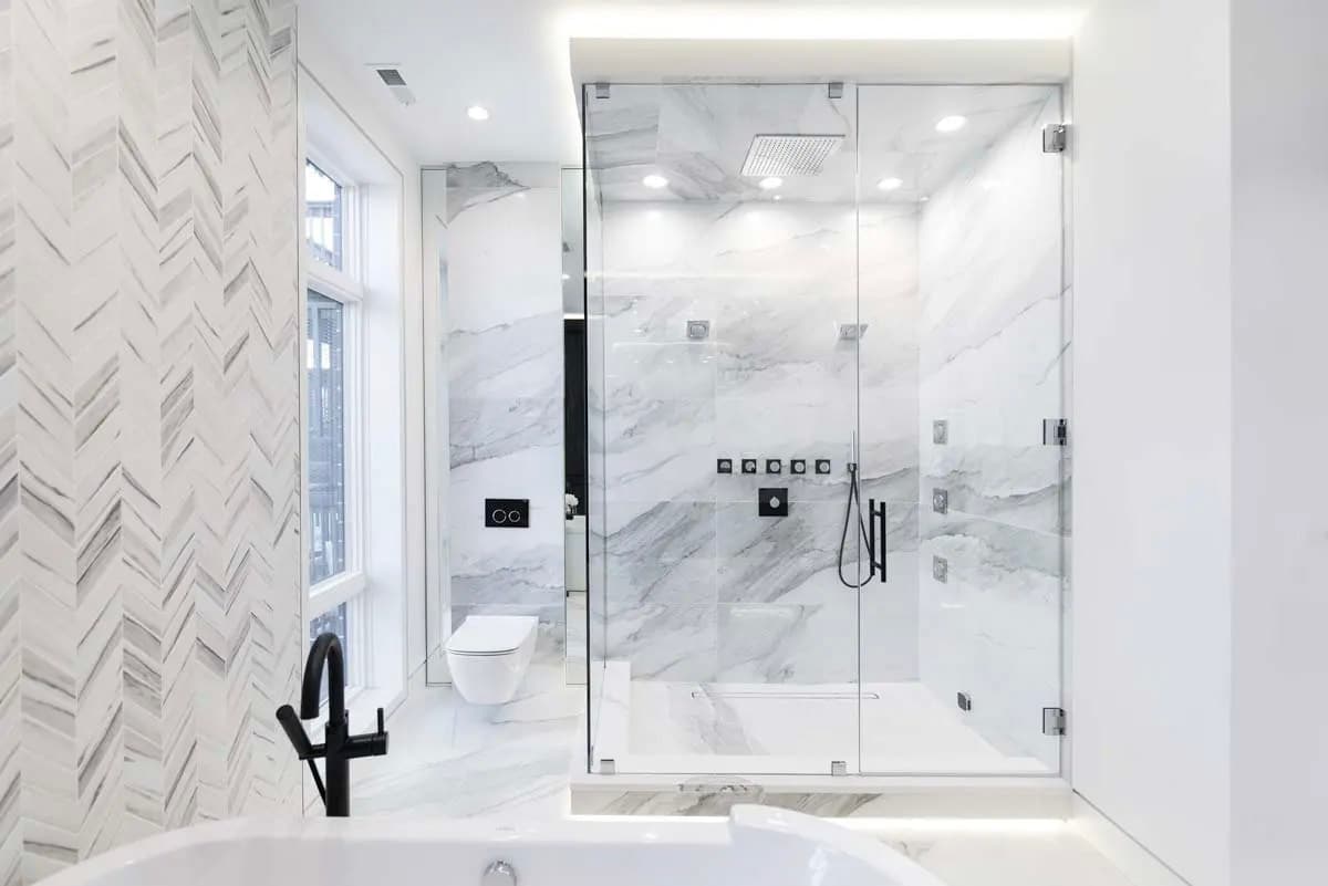 Modern luxury bathroom with glass shower, white marble walls, herringbone tile, black fixtures, white bathtub and recessed lighting for a clean and bright design
