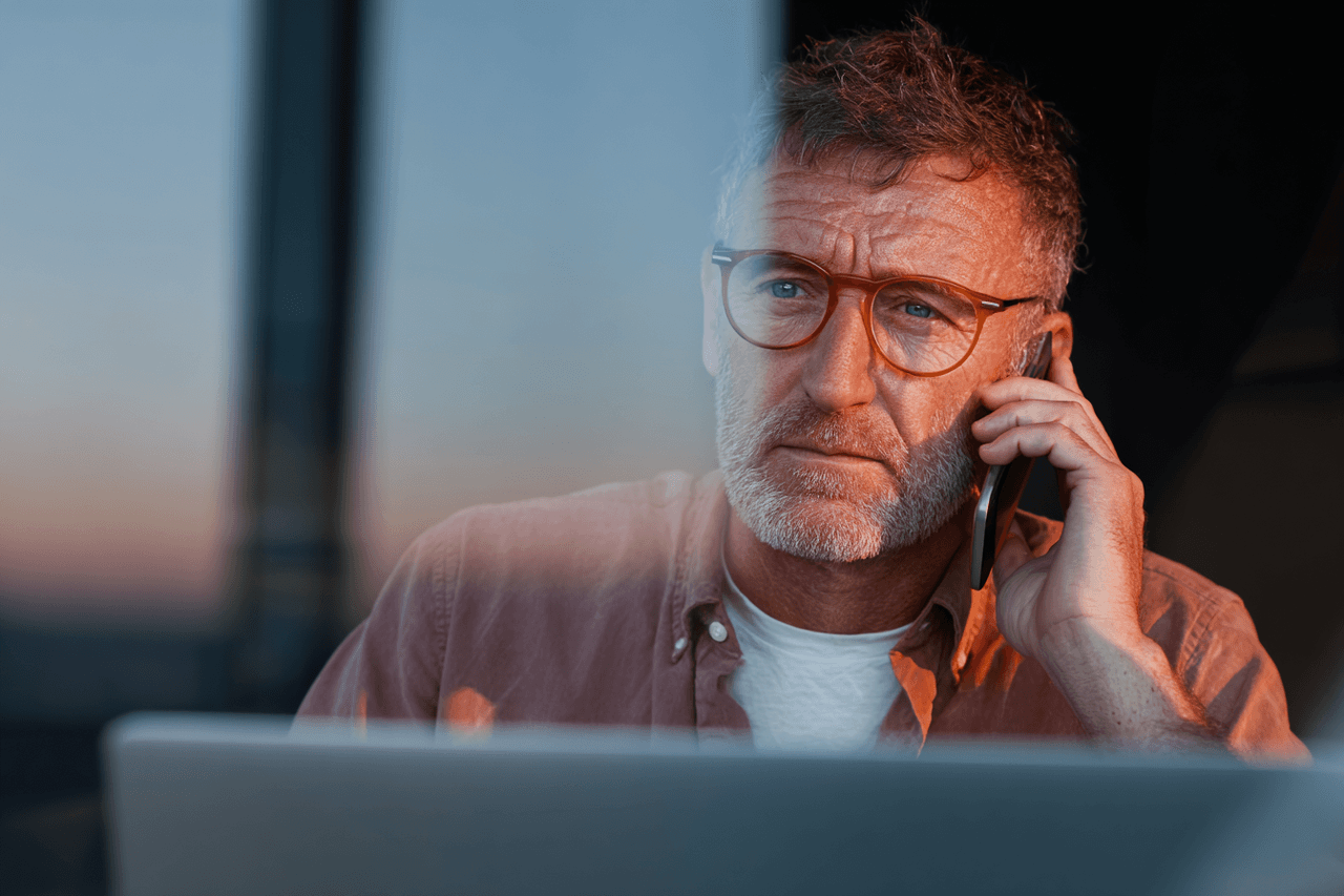 Focused man with glasses talking on the phone in front of a laptop, during sunset.