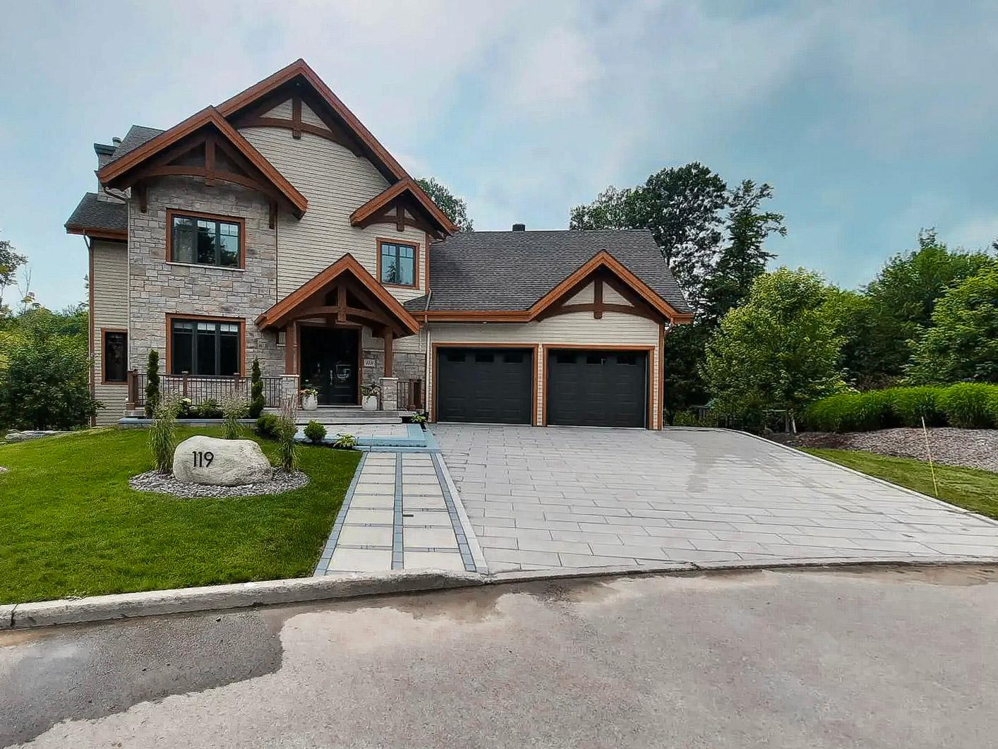 A contemporary two-story stone house with wooden accents on the facade, a large double garage with black doors, a modern paved driveway, and a well-manicured lawn, surrounded by greenery and mature trees.