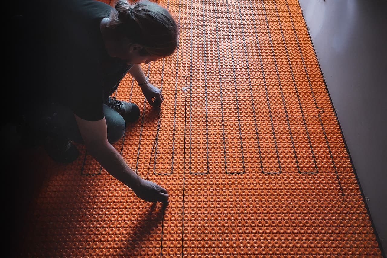 Worker installing an electric underfloor heating system with an orange membrane in a room under construction.