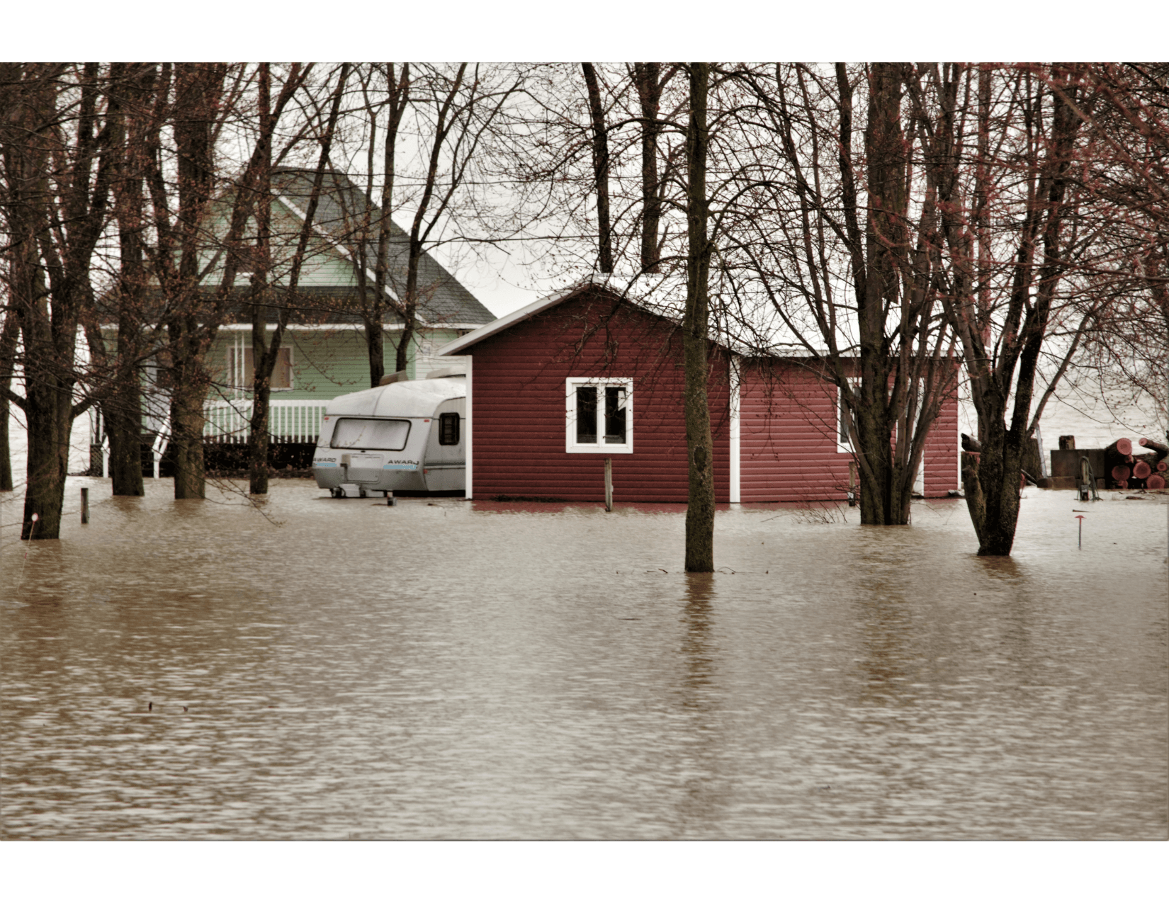 Flooding in a residential area, with a red house surrounded by water and an RV parked nearby. Bare winter trees stand amid the stagnant waters.