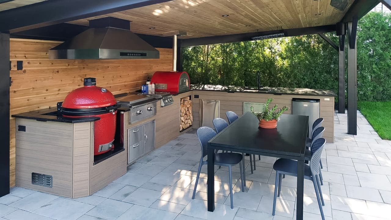 Covered outdoor kitchen with barbecue, smoker, pizza oven, range hood, countertops, built-in storage and dining area on a paved patio.