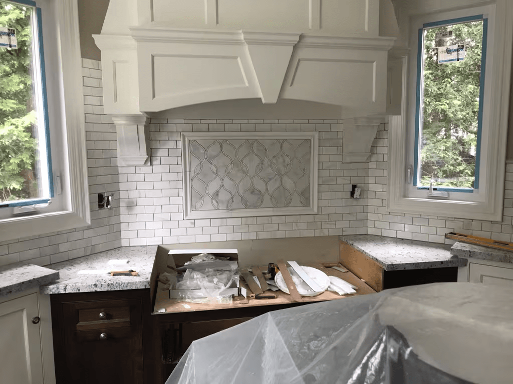 Residential kitchen under renovation with custom cabinetry, white tile backsplash, stone countertops and a decorative range hood being finished