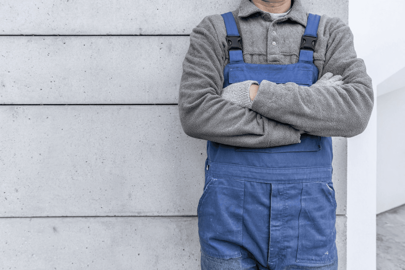 Professional plumber wearing blue overalls and work gloves, standing with arms crossed in front of a concrete wall.