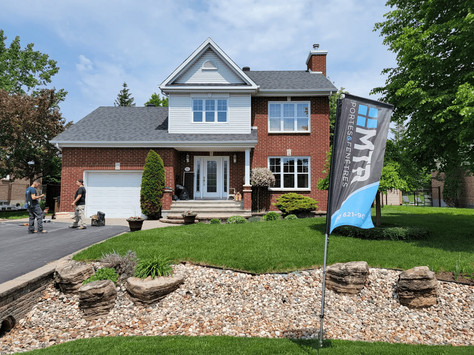 Red brick family home with integrated garage, shingle roof, manicured landscaping, and decorative stone garden bed