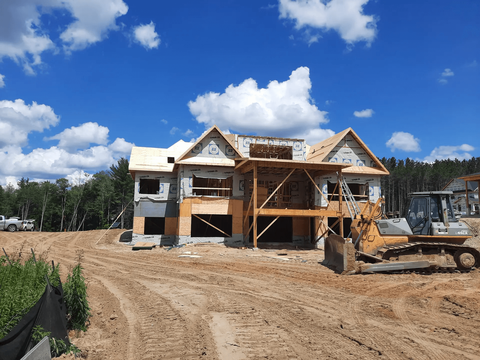 Two-story house under construction on a sandy lot with Tyvek weather wrap, wooden scaffolding, and heavy machinery on site.