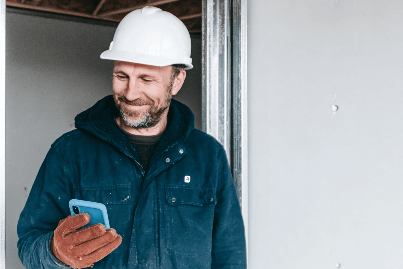 Construction contractor wearing a white hard hat and work gloves while checking his phone inside a house under construction