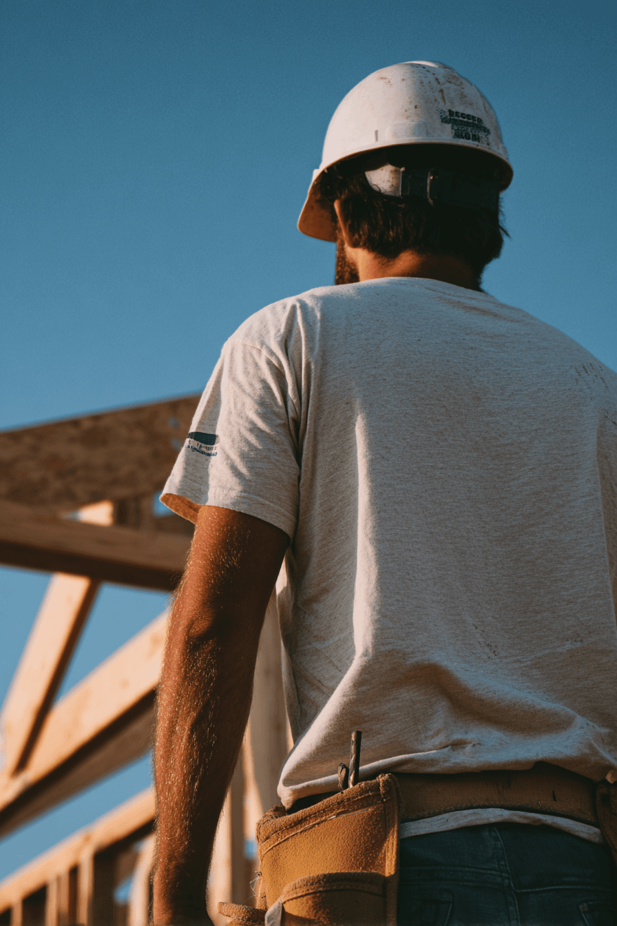 Construction worker seen from behind wearing a hard hat and tool belt in front of wood framing at a job site