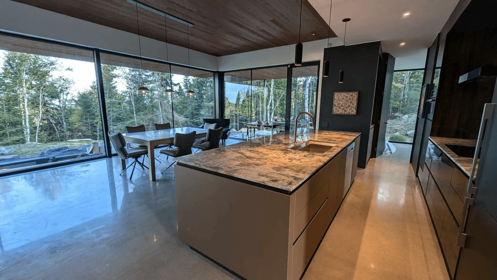 Modern kitchen with a marble island, large glass windows overlooking the forest, wooden ceiling, and polished concrete floor.