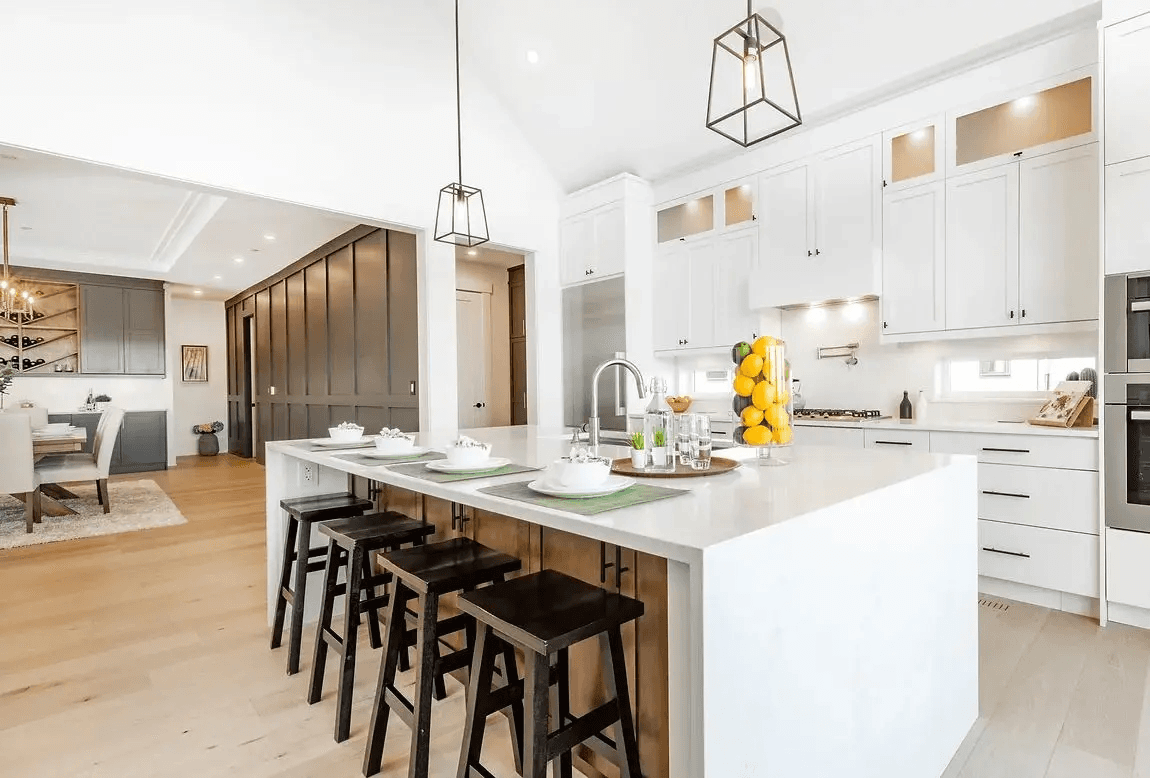 Modern white kitchen with a large central island, black stools, custom cabinetry, pendant lighting and light wood flooring