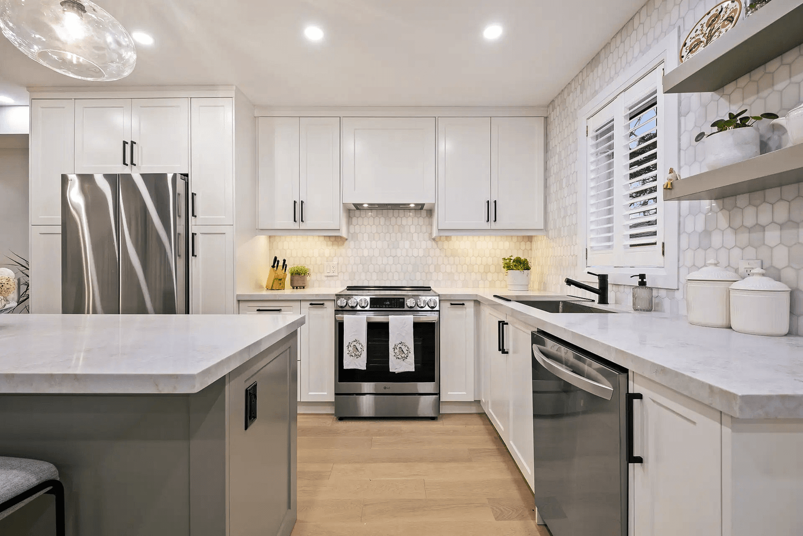 Bright modern kitchen with white cabinets, large central island, light countertops, hexagonal tile backsplash, stainless steel appliances and black sink.